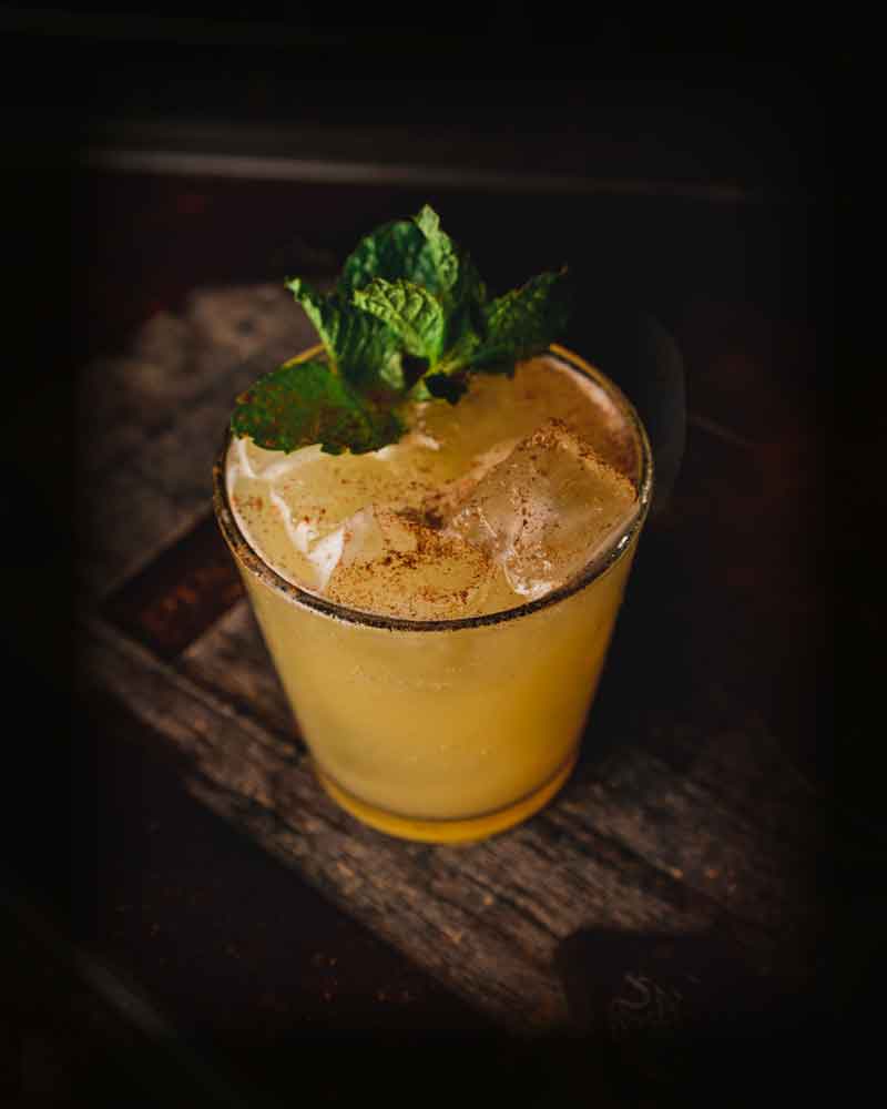 photo of a cocktail sitting on a wooden bar. The drink has is yellow and is garnished with mint leaves.